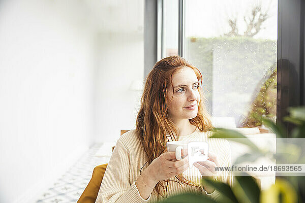 Thoughtful woman sitting with cup in hand by window