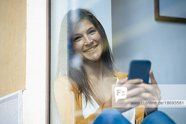 Happy woman holding smart phone in cafe seen through glass window