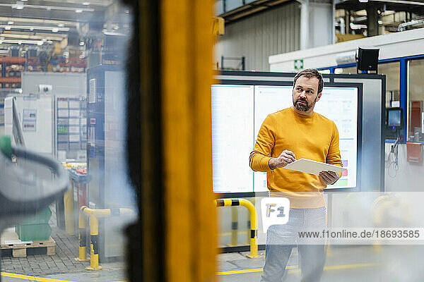 Thoughtful man standing with tablet PC in factory
