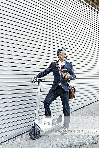Businessman with smart phone and electric scooter standing near wall