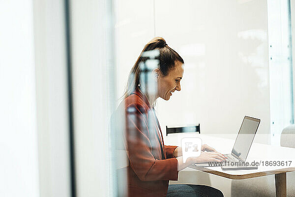 Businesswoman working on laptop sitting at desk seen through glass wall