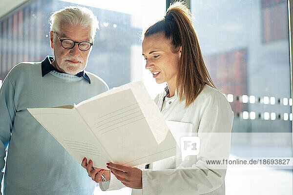 Doctor reading file by senior patient at sunny day in hospital
