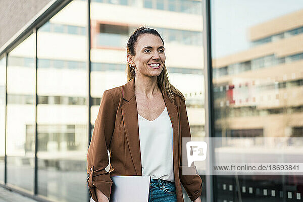 Smiling businesswoman walking near wall holding laptop