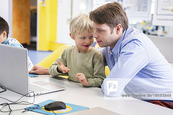 Junge spricht mit einem Lehrer  der in der Schule am Schreibtisch mit Laptop sitzt