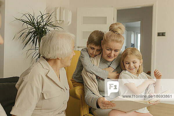 Ältere Frau sitzt mit Tochter und Enkelkindern und schaut sich zu Hause ein Fotoalbum an