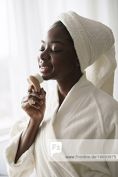 Smiling woman wearing bathrobe using face brush at home