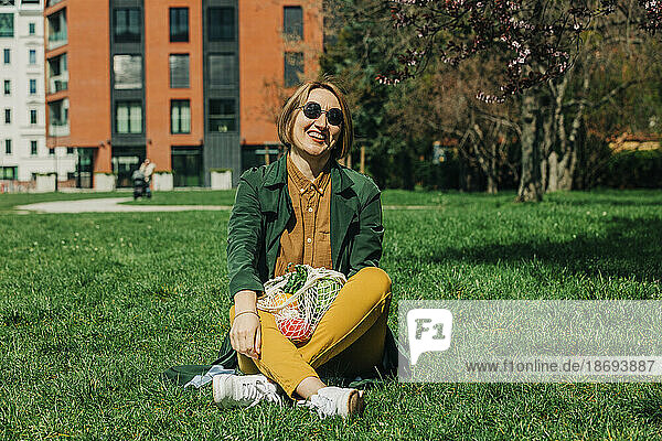 Smiling woman sitting with mesh bag on grass at park