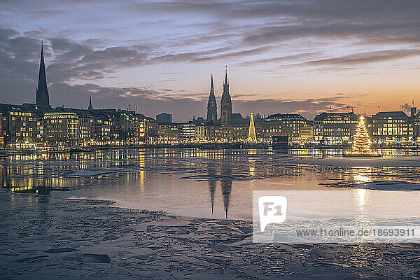 Germany  Hamburg  Ice floating in Alster Lake at dusk with city skyline and glowing Christmas trees in background