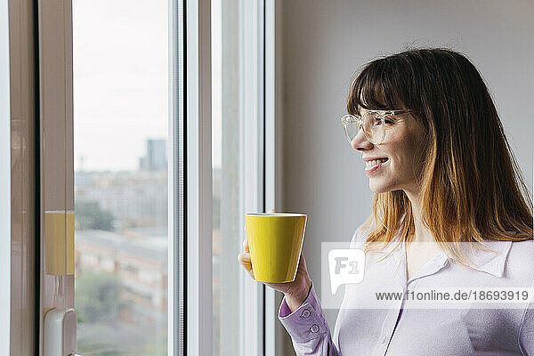 Lächelnde Geschäftsfrau mit Kaffeetasse  die durch das Fenster schaut