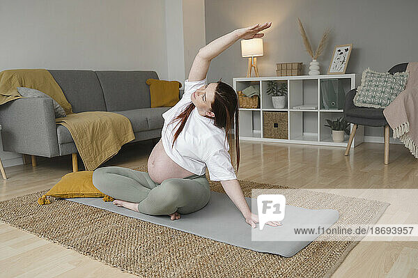 Pregnant woman stretching hand sitting on exercise mat at home