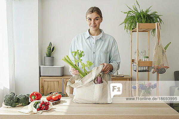 Smiling woman removing celery from reusable bag at home