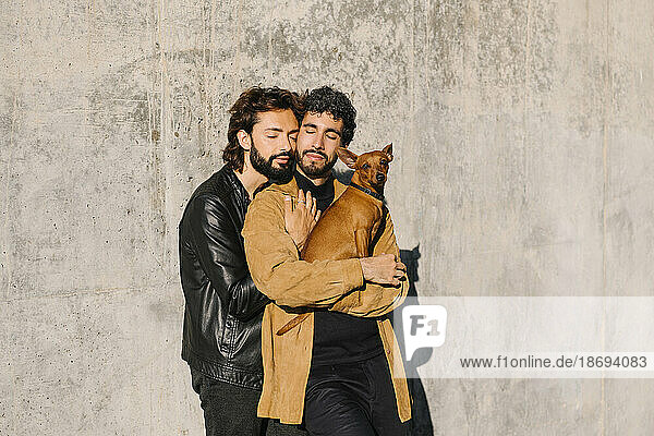 Gay couple with eyes closed carrying dog in front of concrete wall
