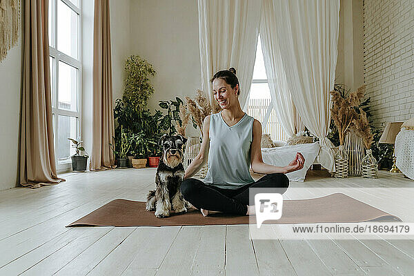 Smiling woman practicing yoga by Schnauzer dog on exercise mat at home