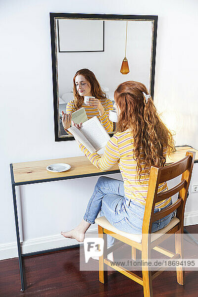 Woman with cup in hand reading book sitting on chair in front of mirror