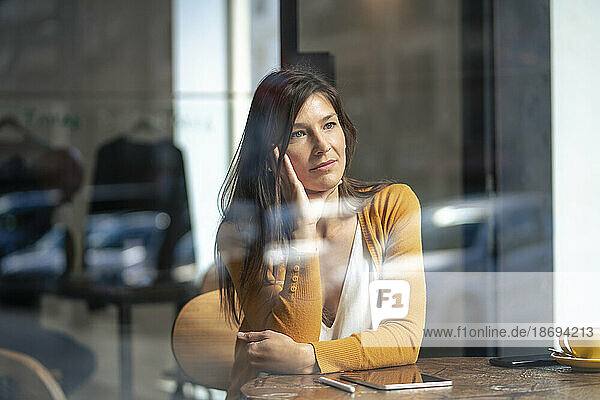 Contemplative woman sitting at table in cafe seen through glass window