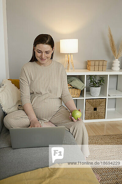 Smiling pregnant freelancer using laptop sitting on sofa at home office