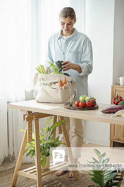 Woman removing organic vegetables from reusable bag standing at home