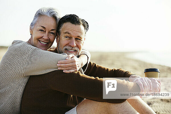 Happy mature woman embracing man holding coffee cup at beach