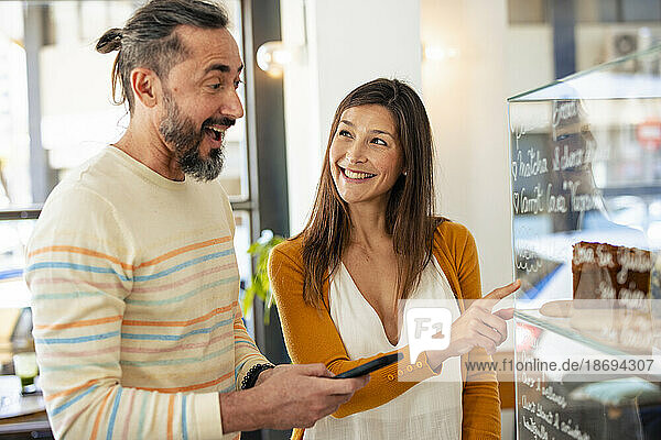 Smiling woman pointing at food display by man in coffee shop