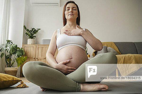 Pregnant woman with eyes closed sitting cross-legged on exercise mat at home