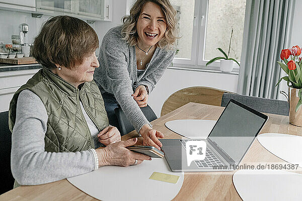 Happy granddaughter by senior woman with laptop on table at home