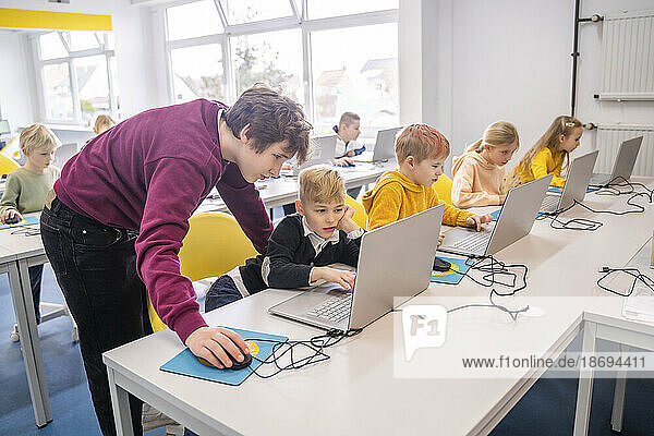 Volunteer assisting boy using laptop sitting at desk with friends in school