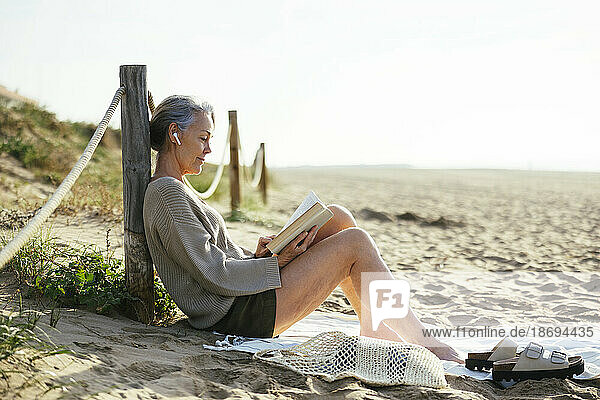 Mature woman wearing wireless in-ear headphones reading book at beach