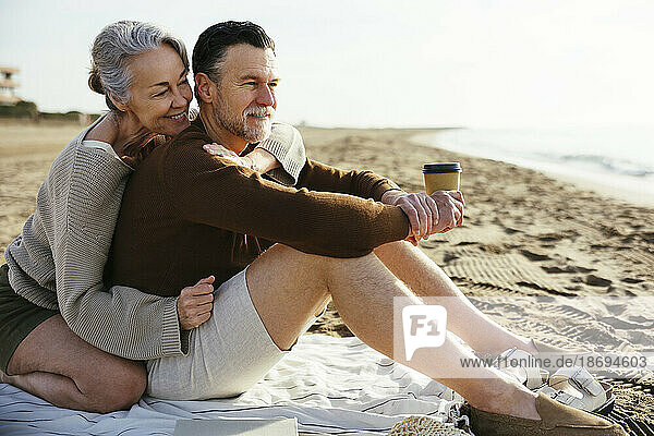 Smiling woman embracing man holding disposable cup at beach