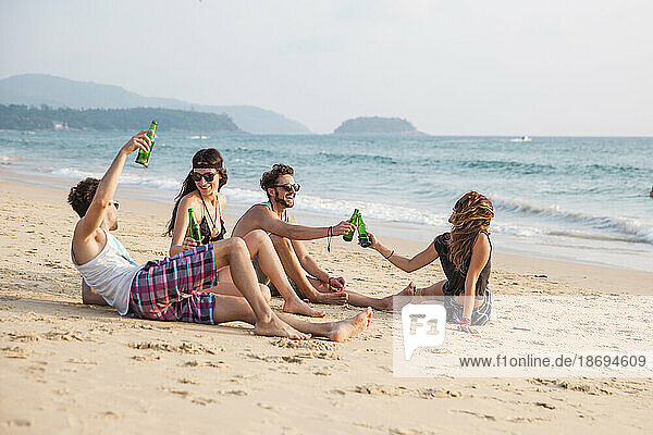 Happy friends toasting drink bottles sitting at beach