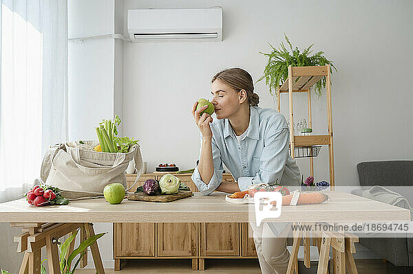 Woman smelling fresh apple leaning on table at home