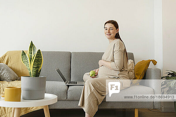 Smiling pregnant freelancer sitting with laptop on sofa at home office