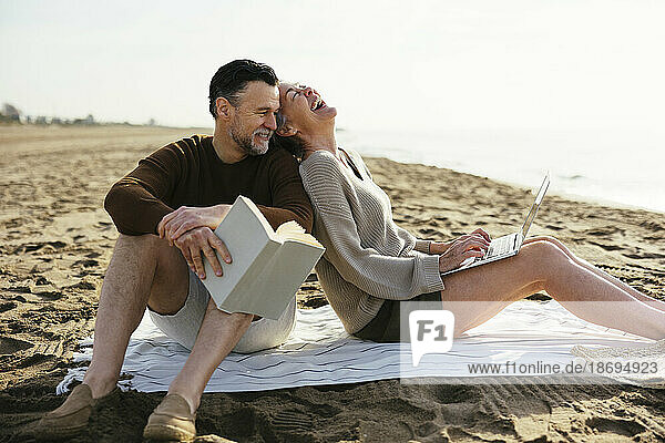 Glückliche Frau mit Laptop sitzt neben Mann und hält Buch am Strand