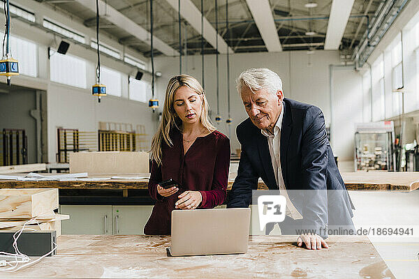 Businesswoman discussing over laptop with businessman leaning on workbench in factory