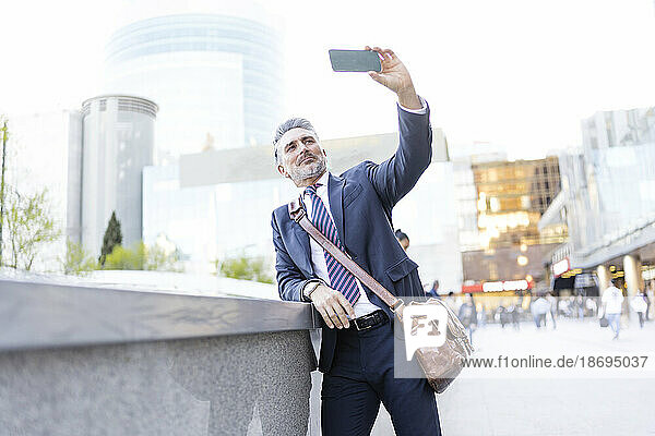 Businessman taking selfie through smart phone leaning on wall