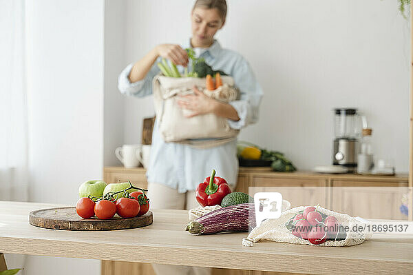 Variety of fresh organic vegetables in reusable bags on table with woman at home