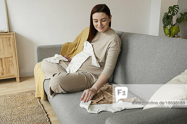 Smiling pregnant woman with baby clothing sitting on sofa at home