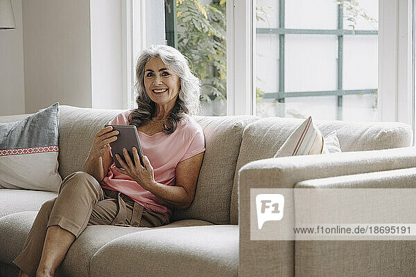 Portrait of smiling mature woman with tablet sitting on couch at home