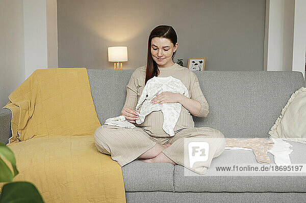 Pregnant woman sitting cross-legged with baby clothing on sofa at home