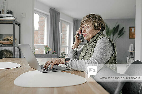 Elderly woman using laptop and talking on smart phone at home