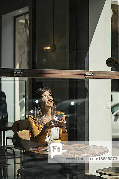 Thoughtful woman with smart phone sitting at cafe seen through glass