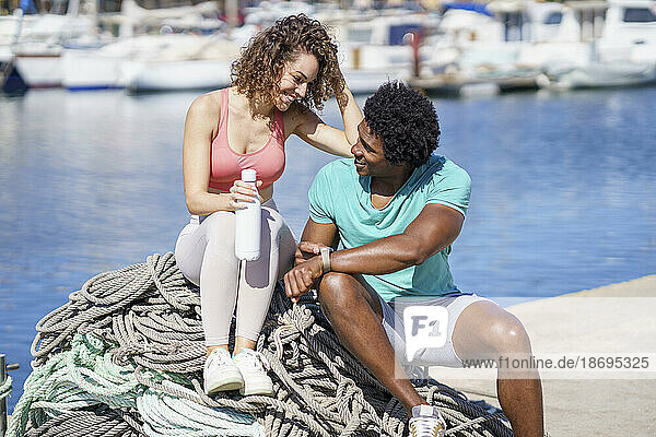 Man showing smart watch to girfriend sitting on rope at harbour