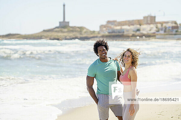 Lächelndes Paar  das mit den Armen am Strand steht