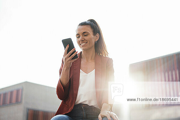 Smiling businesswoman text messaging through smart phone at sunny day
