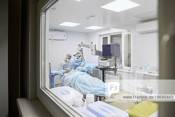 Doctor performing eye surgery in operating room seen through glass window