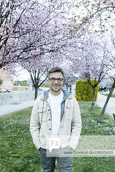 Smiling man standing with hands in pockets near cherry blossom tree at park