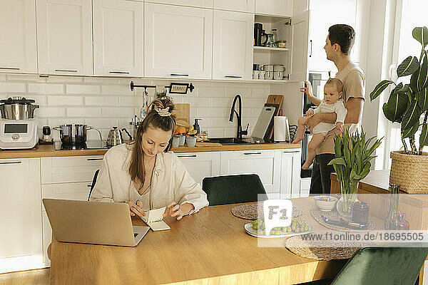 Woman working on laptop with man carrying daughter doing chores in background at home