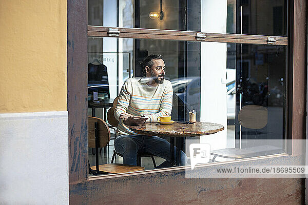 Contemplative man sitting in cafe seen through glass window