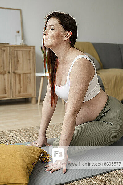 Pregnant woman with eyes closed stretching on mat at home