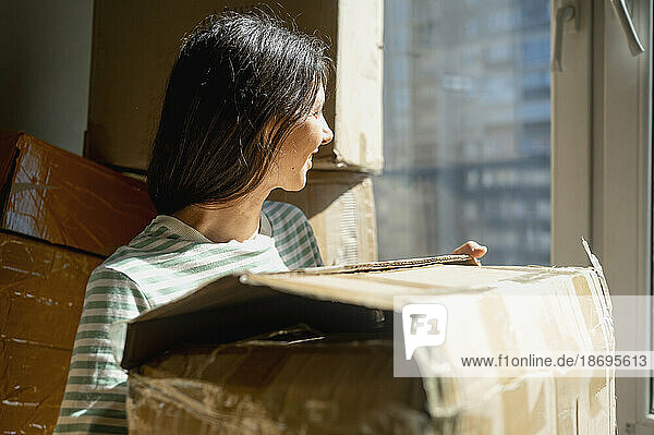 Smiling woman looking out window holding cardboard box at home