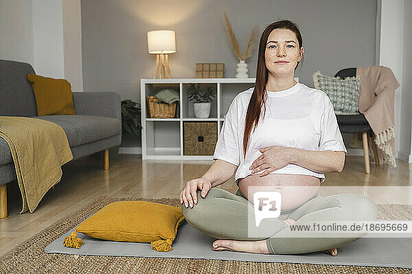 Pregnant woman touching belly sitting cross-legged on mat at home